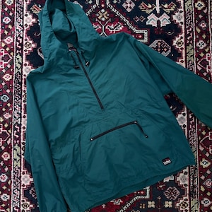 May include: A teal anorak jacket with a hood and a half-zip closure. The jacket has a front pocket with a black zipper and a small logo patch on the lower right. The jacket is made of a lightweight material.