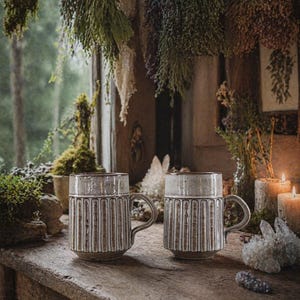 May include: Two ceramic mugs with vertical ribbed designs and a light brown glaze sit on a wooden surface. The mugs have a handle and a contrasting rim. The background features dried plants, candles, and crystals, creating a rustic, bohemian aesthetic.