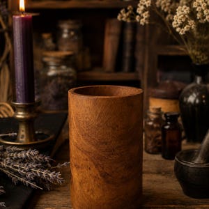 May include: A cylindrical wooden container with a natural wood grain finish. A lit purple candle in a brass holder is to the left. The background includes shelves with jars, bottles, and dried flowers, suggesting a theme of apothecary or natural remedies.