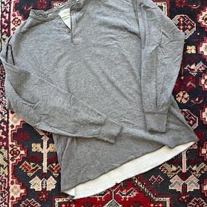 May include: A gray long-sleeved Henley shirt with a three-button placket. The shirt has a crew neck and a white lining visible at the bottom hem. The shirt is made of a soft material and is laid flat on a patterned rug.