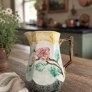 May include: A ceramic pitcher featuring a raised floral design with pink flowers and green leaves on a light blue background. The top rim is yellow with a textured pattern, and the base has a dark gray, intricate relief design. It has a brown handle.