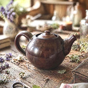 May include: A brown ceramic teapot with a lid and handle sits on a rustic wooden table. The teapot is surrounded by dried flowers, a key, and a small book. The scene evokes a vintage, cozy aesthetic.
