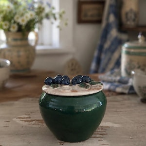 May include: A dark green ceramic jar with a rounded body and a matching lid. The lid features a sculpted cluster of dark blue blueberries with green leaves attached to the top surface. The jar has a smooth, glossy finish and a wide, circular base.