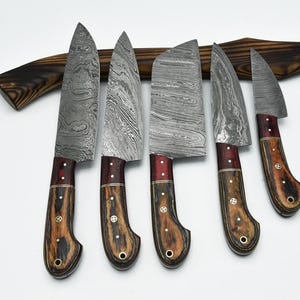 May include: A set of five knives with patterned Damascus steel blades and wooden handles featuring dark grain and red accents. The handles are secured with metal rivets and include a lanyard hole at the base. A curved wooden handle rests behind.