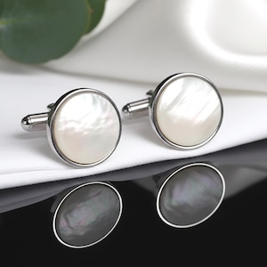 Könnte beinhalten: Zwei silberne Manschettenknöpfe mit einem runden Perlmutt-Design. Sie werden auf weißem Satin präsentiert, mit einer dunklen, reflektierenden Oberfläche darunter. Grüne Eukalyptusblätter sind zu sehen.