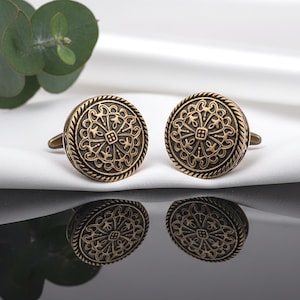 Peut inclure: Une paire de boutons de manchette dorés antiques. Chaque bouton de manchette présente un motif floral complexe dans un cercle avec une bordure en forme de corde. Ils sont présentés sur une surface blanche avec une base noire réfléchissante.