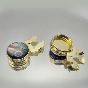 以下が含まれることがあります： 2つのゴールドカラーのカフスボタンセット。1つのカフスボタンは、ゴールドカラーの金属ベースにセットされた、丸く虹色の濃い色の石を持っています。もう一方のカフスボタンは開いており、ゴールドカラーの金属ベースが明らかになっています。