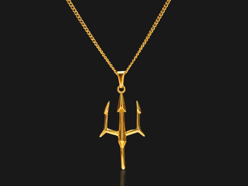 Puede incluir: Un collar con colgante dorado con dise&ntilde;o de tridente. El tridente, una lanza de tres puntas, cuelga de una delicada cadena. La joya se presenta sobre un fondo oscuro, resaltando el brillo met&aacute;lico del colgante.