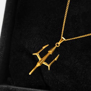 Puede incluir: Un collar con colgante de tridente dorado. El colgante, un dise&ntilde;o de lanza de tres puntas, cuelga de una fina cadena. El collar se muestra sobre un fondo negro, resaltando los detalles de la joya.