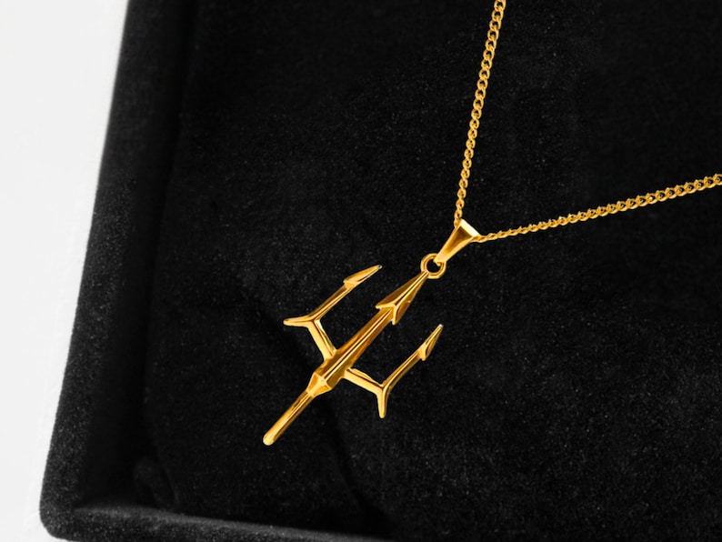 Puede incluir: Un collar con colgante de tridente dorado. El colgante, un dise&ntilde;o de lanza de tres puntas, cuelga de una fina cadena. El collar se muestra sobre un fondo negro, resaltando los detalles de la joya.