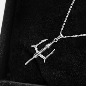 Puede incluir: Un collar con colgante plateado con dise&ntilde;o de tridente. El colgante cuelga de una fina cadena y descansa sobre un fondo de terciopelo negro. El tridente tiene tres puntas y una lanza central.