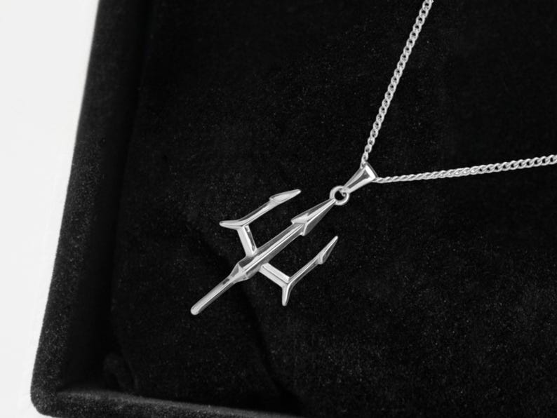 Puede incluir: Un collar con colgante plateado con dise&ntilde;o de tridente. El colgante cuelga de una fina cadena y descansa sobre un fondo de terciopelo negro. El tridente tiene tres puntas y una lanza central.