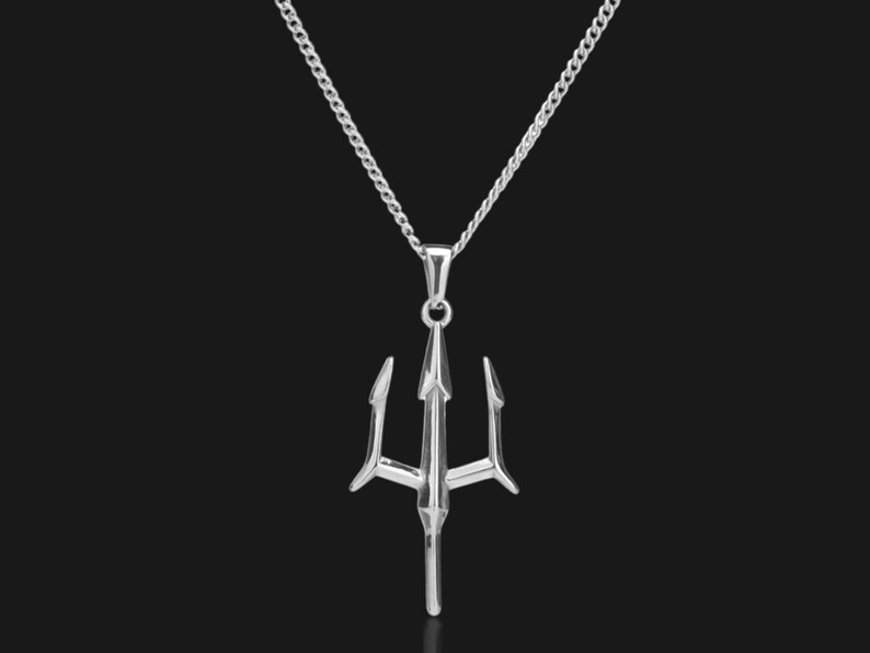 Puede incluir: Un collar con colgante plateado con un dise&ntilde;o de tridente. El tridente, una lanza de tres puntas, cuelga de una delicada cadena. El collar se muestra sobre un fondo oscuro, resaltando el metal brillante del colgante y la cadena.