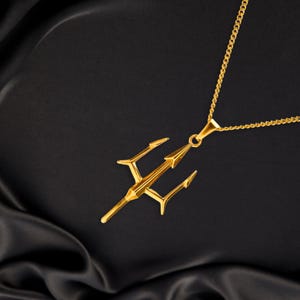 Puede incluir: Un collar con colgante dorado con un dise&ntilde;o de tridente. El tridente tiene tres puntas y una punta de lanza central, suspendida de una delicada cadena. El collar se muestra sobre un fondo negro y sedoso.
