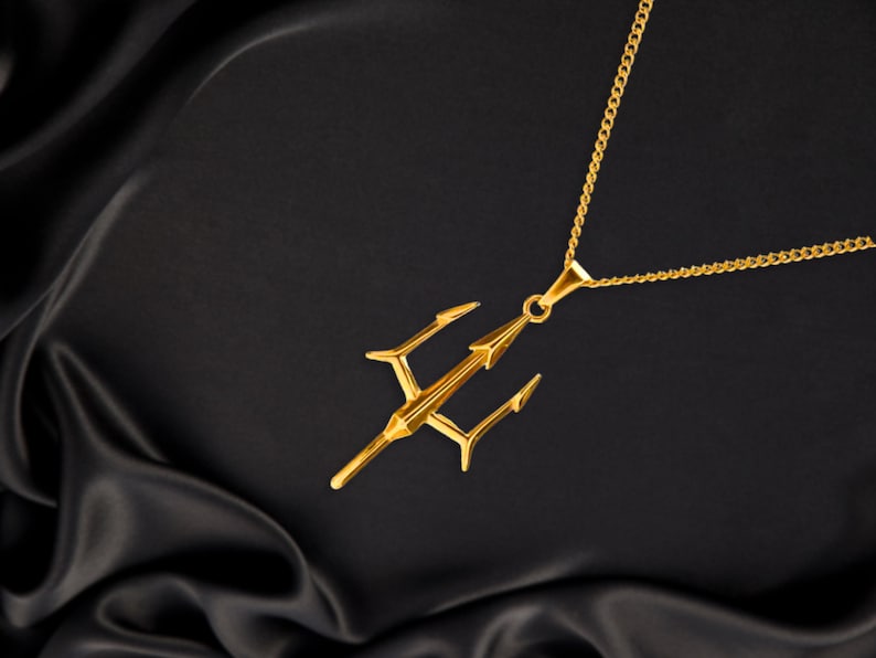Puede incluir: Un collar con colgante dorado con un dise&ntilde;o de tridente. El tridente tiene tres puntas y una punta de lanza central, suspendida de una delicada cadena. El collar se muestra sobre un fondo negro y sedoso.