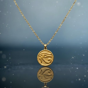 Puede incluir: Un collar dorado con un colgante circular con el símbolo del Ojo de Horus. El colgante y la cadena son de un material metálico brillante. El collar cuelga sobre un fondo azul oscuro borroso.