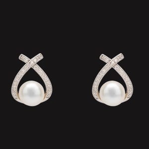以下が含まれることがあります： 白い真珠を、キラキラ光るシルバーカラーの交差したフレームで囲んだ、エレガントなイヤリングのペア。デザインは暗い背景に配置され、ジュエリーの繊細なディテールを際立たせています。