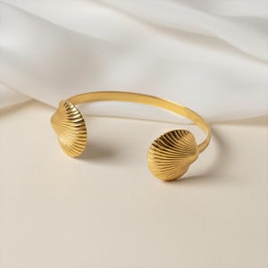 Könnte beinhalten: Ein goldfarbenes Armreif-Armband mit Muscheldesign. Das Armband hat zwei muschelförmige Enden mit detaillierten geriffelten Texturen. Das offene Design ermöglicht ein einfaches Tragen. Das Armband wird auf einer weißen Oberfläche präsentiert.