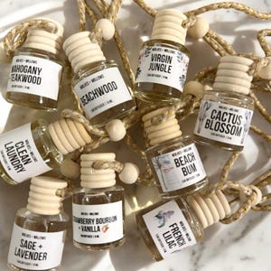 Puede incluir: Una colección de ambientadores para coche en pequeños frascos de vidrio con tapas de madera. Cada frasco tiene una etiqueta con un nombre de aroma diferente, como "Mahogany Teakwood" y "Cactus Blossom". Los frascos están conectados por una cuerda dorada.