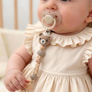 Puede incluir: Un bebé con un chupete y un clip de chupete de madera. El clip presenta una cabeza de oso de ganchillo, cuentas de madera y bloques de letras de madera que deletrean "Kylie". El chupete tiene una tetina de silicona beige. Una estrella de madera está unida al extremo.