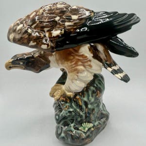 May include: A ceramic figurine of an eagle in flight, perched atop a textured, mottled green and brown rock base. The eagle features glossy brown, white, and black plumage, with its wings extended horizontally and its head turned to the left.