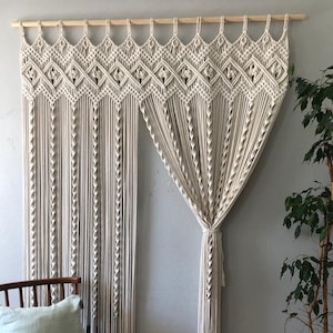 May include: Cream-colored macrame curtain with geometric patterns and long, vertical fringe. The curtain is suspended from a wooden rod, adding a bohemian touch to the room. The design features intricate knotting and a textured appearance.