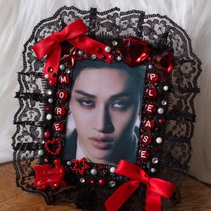 May include: A framed portrait with a black lace border, red bows, and decorative red gems. The frame features the words "MORE PLEASE" spelled out with letter beads. The portrait is of a person with dark hair and a serious expression.