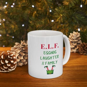 May include: White ceramic mug with the text "E.L.F. EGGNOG LAUGHTER & FAMILY" in red and green. The mug features a green elf hat with red and yellow striped legs. The mug is on a wooden surface with pinecones in the background.