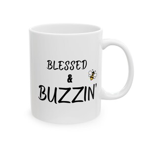Puede incluir: Taza de cerámica blanca con las palabras "BLESSED & BUZZIN'" en escritura negra. A la derecha del texto hay una pequeña abeja de dibujos animados con rayas amarillas y negras. La taza tiene un asa curva.