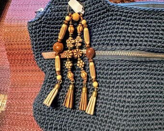 Puede incluir: Un bolso de mano de ganchillo azul marino con una correa trenzada marrón y un adorno de borla de madera decorativa. La borla presenta cuentas de madera, detalles florales y pequeñas borlas beige. El bolso tiene un cierre de cremallera.