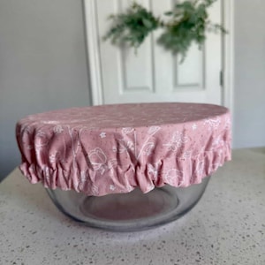 May include: A clear glass bowl covered with a pink fabric lid. The lid has a white floral pattern and a ruffled edge. The bowl is sitting on a white countertop.
