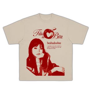 Pode incluir: Uma t-shirt bege de manga curta com uma impressão gráfica vermelha de uma pessoa, o texto "Take a Bite", "beabadoobee" e "i wanted to see the world in colour // through your eyes and through your mind" com uma ilustração de maçã.