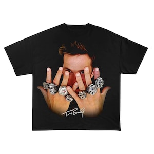 Könnte beinhalten: Schwarzes T-Shirt mit einer Grafik einer Person mit mehreren Ringen an den Fingern, wobei die Augen der Person sichtbar sind. Die Hände der Person sind erhoben und zeigen die Ringe. Die Unterschrift "Tom Brady" befindet sich unten.