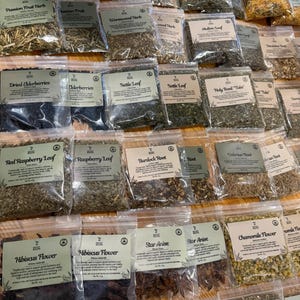 Puede incluir: Una colecci&oacute;n de bolsas de pl&aacute;stico transparente llenas de diversas hierbas, ra&iacute;ces y flores secas, cada una con una etiqueta de Seven Roots que muestra el nombre del producto y una breve descripci&oacute;n de sus usos.