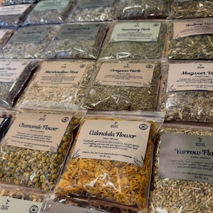 May include: A display of various dried herbs in clear plastic bags. The bags are labeled with names like "Rosemary Herb," "Oregano Herb," and "Calendula Flower." The herbs are arranged on a wooden surface, showcasing a variety of colors and textures.