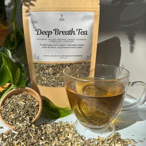 Puede incluir: Una taza de té de vidrio transparente con un infusor de metal, llena de té. Una bolsa de "Deep Breath Tea" está detrás de la taza, junto con un pequeño cuenco de madera y hojas de té sueltas. La bolsa de té enumera los ingredientes e instrucciones.