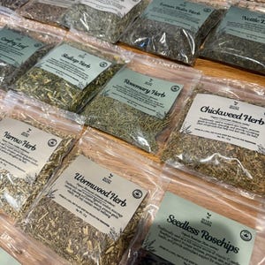 Puede incluir: Una colecci&oacute;n de bolsas de pl&aacute;stico transparente llenas de diversas hierbas y productos bot&aacute;nicos secos, cada una con una etiqueta blanca con el logotipo de Seven Roots, el nombre del producto y texto descriptivo. Las hierbas incluyen romero, pamplina, ajenjo y escaramujos.