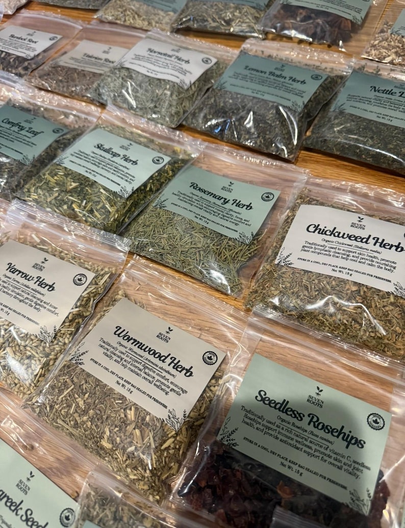 Puede incluir: Una colecci&oacute;n de bolsas de pl&aacute;stico transparente llenas de diversas hierbas y productos bot&aacute;nicos secos, cada una con una etiqueta blanca con el logotipo de Seven Roots, el nombre del producto y texto descriptivo. Las hierbas incluyen romero, pamplina, ajenjo y escaramujos.