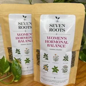 Puede incluir: Dos bolsas de papel marrón con la mezcla de hierbas Seven Roots Women's Hormonal Balance. Las etiquetas blancas muestran un logotipo de planta, texto e ilustraciones de hoja de frambuesa, manzanilla, albahaca sagrada, damiana, hierbabuena y milenrama. 27 g.