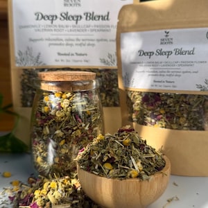Peut inclure: Plusieurs produits de tisane à base de plantes, dont un bocal en verre et un bol en bois remplis d'un mélange d'herbes et de fleurs séchées. Deux sacs en papier brun avec le texte "Deep Sleep Blend" sont visibles. Le mélange comprend de la camomille, de la mélisse et de la lavande.