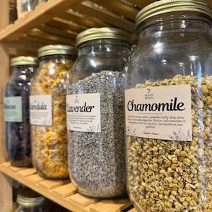 May include: A shelf displays glass jars filled with dried herbs and flowers. The jars have gold lids and white labels with text. The labels read "Chamomile" and "Lavender". The jars are arranged on wooden shelves.