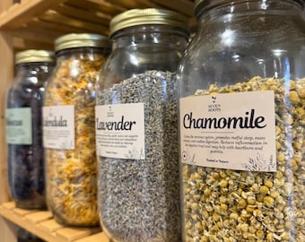 Peut inclure: Des bocaux en verre remplis d'herbes et de fleurs séchées sont disposés sur une étagère en bois. Les bocaux ont des couvercles dorés et des étiquettes blanches. Les étiquettes indiquent "Camomille" et "Lavande".