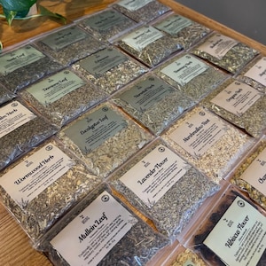 May include: An assortment of dried herbs in clear plastic bags, each labeled with the herb's name. The bags are arranged on a wooden tray. Herbs include Damiana Leaf, Eucalyptus Leaf, and Lavender Flower.