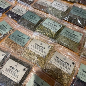 Puede incluir: Una colecci&oacute;n de bolsas de pl&aacute;stico transparente llenas de diversas hierbas y ra&iacute;ces secas, cada una con una etiqueta verde claro con el logotipo de Seven Roots, el nombre del producto y el peso neto en gramos. El surtido incluye Mullein Leaf, Yarrow Herb y Wormwood Herb.