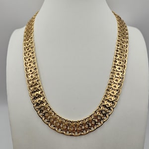 May include: A gold-toned necklace with a wide, intricate chain design. The necklace is displayed on a white mannequin bust. The chain features a repeating pattern of openwork links, creating a textured and eye-catching appearance. The necklace is a statement piece.
