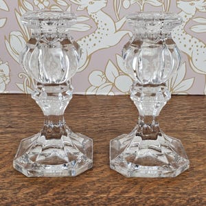May include: Two clear glass candle holders with a faceted base and a bulbous, ribbed centre section. The candle holders are displayed on a wooden surface, with a floral and deer-patterned wallpaper in the background.