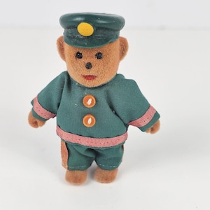 May include: A small, plush teddy bear figurine dressed in a green uniform with a matching cap. The uniform has pink trim and orange buttons. The bear has a brown face and is standing upright.