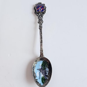 Puede incluir: Una cuchara de plata vintage con un diseño de Disneyland. El cuenco de la cuchara muestra una imagen colorida de un castillo contra un cielo azul. El mango tiene un diseño retorcido y un escudo de Disneyland.