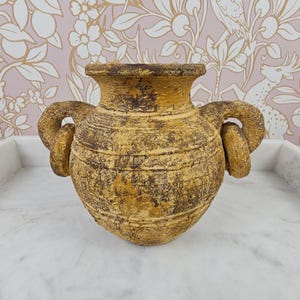 Può includere: Un vaso in ceramica color giallo senape di forma rotonda, con una finitura strutturata e invecchiata e accenti marrone scuro. Il recipiente presenta un bordo largo e svasato e due manici circolari spessi e prominenti sui lati.