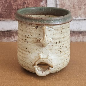 Könnte beinhalten: Handgefertigter Keramik-Gesichtstopf mit gesprenkelter, cremefarbener Oberfläche. Der Topf hat eine modellierte Nase und einen offenen Mund mit grünem Rand. Ein einzigartiges, künstlerisches Keramikstück für die Heimdekoration oder als Gebrauchsgegenstand.